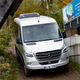 a silver 2023 mercedes benz sprinter awd van navigates a narrow muddy trail down a steep hil between a concrete wall and a stand of trees.